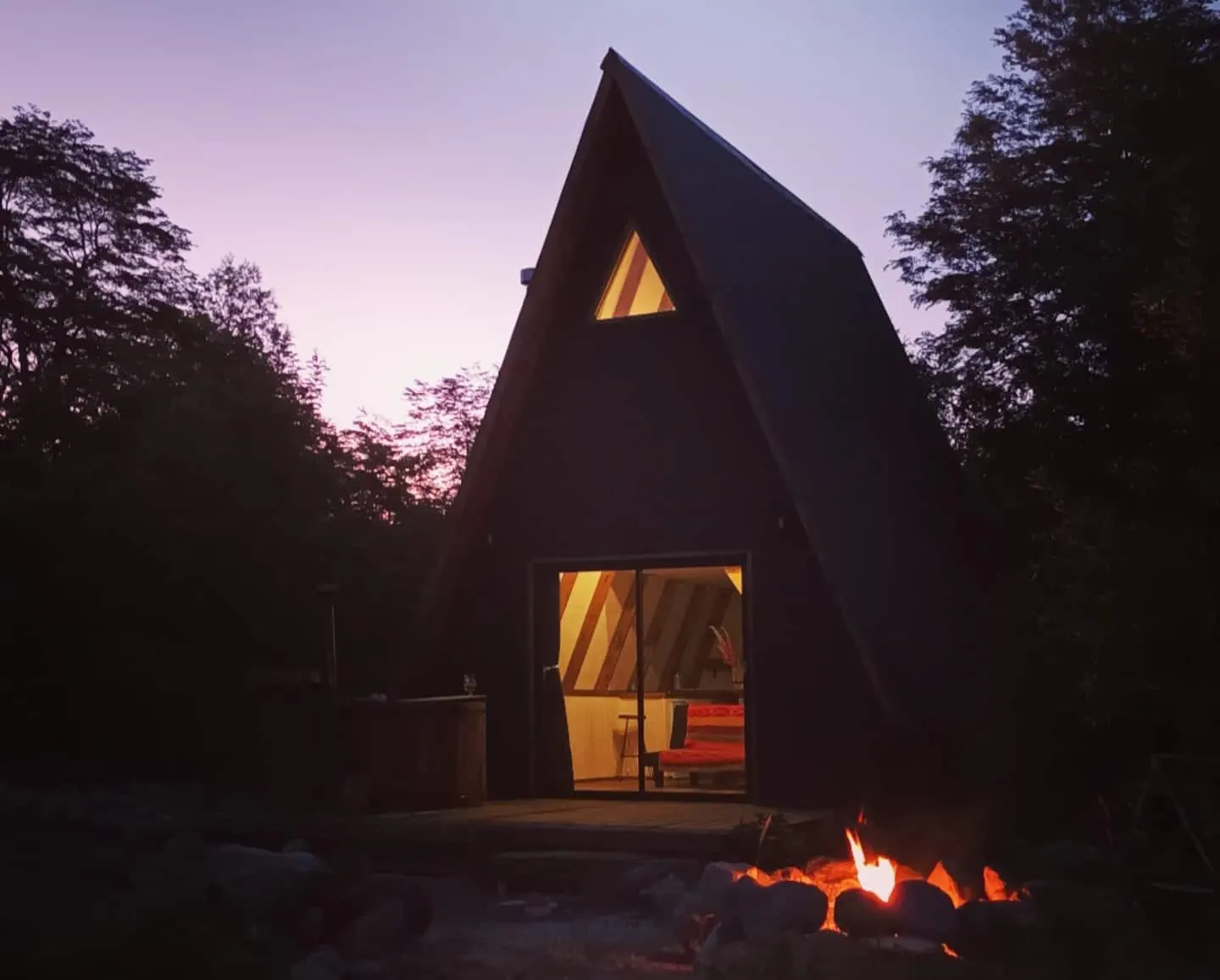 Le chalet en A de Molco Alpina à la tombée de la nuit, fenêtre éclairée et brasero allumé