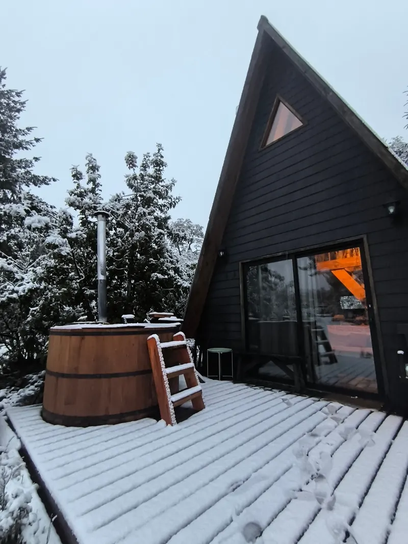 The wooden hot tub and the deck covered in snow, next to the cabin with warm light inside